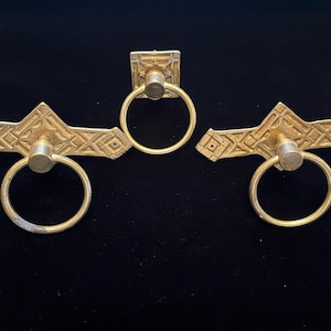 May include: Three brass-toned cabinet pulls and a knob. The pulls have a geometric design and a ring handle. The knob is square with a ring handle. All pieces are on a black background.