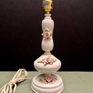 May include: A white ceramic table lamp with a decorative floral design. The lamp features pink roses and green leaves. The lamp has a gold-colored bulb socket and a white cord. The lamp is approximately 15 inches tall.