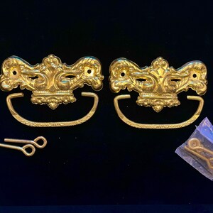 May include: Two ornate gold-colored drawer pulls with matching handles and hardware. The pulls have a decorative floral design and are accompanied by screws, nuts, and additional hardware in a small bag. Ideal for furniture restoration.