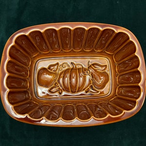 May include: A brown ceramic baking dish with a fluted rim and a raised design of a pumpkin and two pears in the center.
