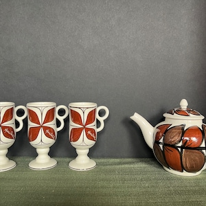 May include: A vintage tea set featuring three stemmed mugs and a teapot. The mugs and teapot are white with a brown floral design. The mugs have handles and the teapot has a lid and spout.