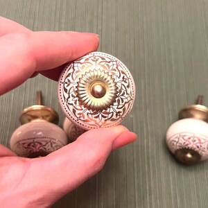 May include: White and gold decorative cabinet knobs. The knobs have a round shape with a gold floral design and a brass base. The knobs are held in a hand against a green background.