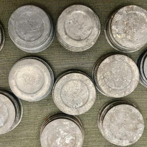 May include: A collection of weathered, round, metal jar lids. The lids are a dull gray color and some have the word "ATLAS" embossed on them. The lids are stacked in small piles.