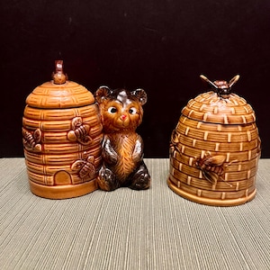 May include: A ceramic honey pot set featuring two beehive-shaped jars with lids and a small bear figurine. The jars are brown with bee and honeycomb details. The bear is brown with a red nose.