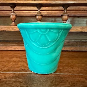 May include: A vibrant turquoise ceramic planter featuring a raised, repeating scalloped pattern around the exterior. The vessel has a wide, circular opening that tapers down to a narrower base, showcasing a smooth, glossy finish throughout.