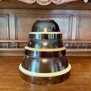 May include: A set of three stacked nesting bowls with a dark brown glazed exterior and a contrasting cream-colored band around the rim of each bowl. The bowls are circular with smooth, rounded sides and a glossy finish, arranged from smallest to largest.