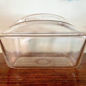 May include: A clear, rectangular glass baking dish with a matching lid. The lid has a curved handle. The dish is sitting on a wooden surface. The dish is designed for baking and food storage.