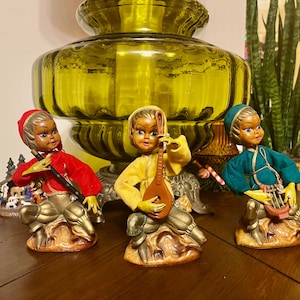May include: Three vintage figurines of musicians. Each figure is dressed in colorful clothing and playing a different instrument. The figurines are on a brown surface, with a large, yellow glass vase in the background.