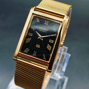May include: A gold-toned Seiko Quartz watch with a rectangular black face and gold Roman numerals. The watch features a mesh band and is displayed on a clear stand. The watch face has the Seiko Quartz logo.