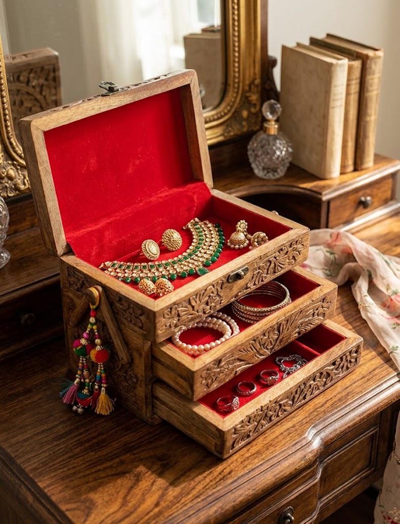 May include: A wooden jewelry box with intricate carvings, featuring a hinged lid and two pull-out drawers. The interior is lined with bright red velvet, holding a gold-tone necklace, earrings, bracelets, and rings. A colorful tassel hangs from the side.