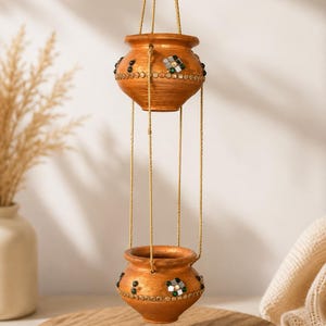 May include: A two-tiered hanging planter set featuring terracotta-colored clay pots suspended by thin golden cords. Each pot is decorated with small, reflective mirror pieces and dark green beads arranged in a circular pattern around the exterior.