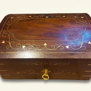 May include: A rectangular wooden box with a dark brown finish and a curved lid. The top and front feature delicate gold-colored metal inlay in floral and vine patterns. A small gold-colored metal latch is centered on the front panel.
