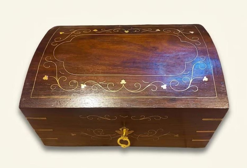 May include: A rectangular wooden box with a dark brown finish and a curved lid. The top and front feature delicate gold-colored metal inlay in floral and vine patterns. A small gold-colored metal latch is centered on the front panel.