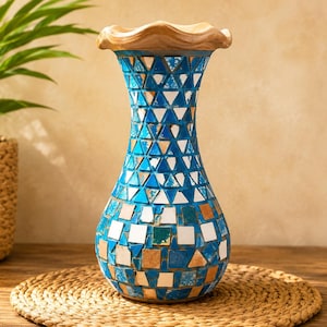 Op de afbeelding: Een decoratieve vaas met een mozaïekpatroon van blauwe, witte en beige tegels. De vaas heeft een smalle hals en een brede, ronde basis met een golvende, lichtgekleurde houten rand aan de bovenkant. Het oppervlak is bedekt met kleine, geometrische stukjes glas.
