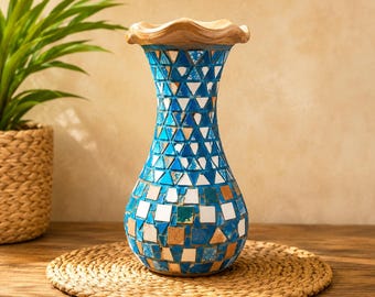 May include: A decorative vase featuring a mosaic pattern of blue, white, and tan tiles. The vase has a narrow neck and a wide, rounded base with a wavy, light-colored wooden rim at the top. The surface is covered in small, geometric glass pieces.
