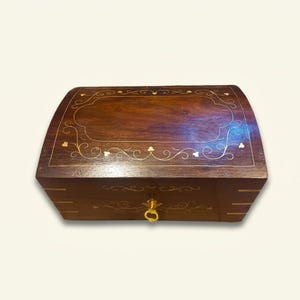 May include: A rectangular dark brown wooden box with a curved lid featuring gold-colored metal inlay in a delicate floral and vine pattern. The front of the box includes a small gold-colored metal latch centered on the front panel.