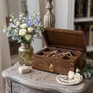 May include: A rectangular dark wood jewelry box with a hinged lid, gold-toned hardware, and delicate gold filigree detailing. The box contains multiple compartments holding pearl necklaces and rings, with a ring box and gold chain nearby.