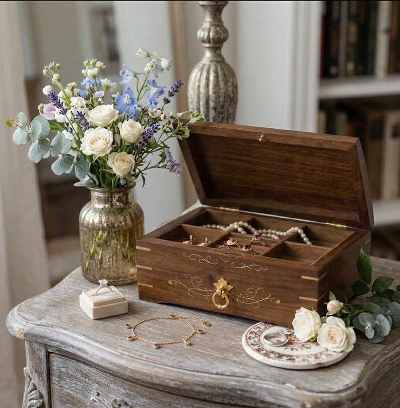 May include: A rectangular dark wood jewelry box with a hinged lid, gold-toned hardware, and delicate gold filigree detailing. The box contains multiple compartments holding pearl necklaces and rings, with a ring box and gold chain nearby.