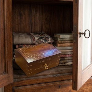May include: A rectangular dark wood box with a hinged lid featuring intricate gold-colored inlay patterns and a small brass ring pull handle. The box is positioned inside a wooden cabinet next to a stack of folded textiles and books.
