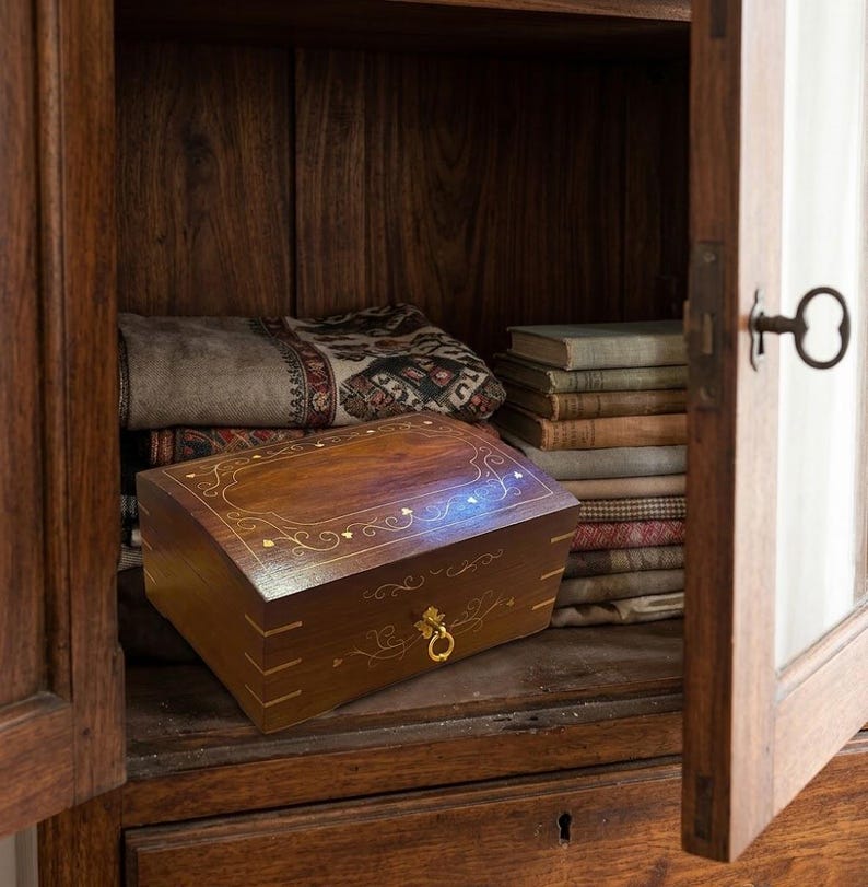 May include: A rectangular dark wood box with a hinged lid featuring intricate gold-colored inlay patterns and a small brass ring pull handle. The box is positioned inside a wooden cabinet next to a stack of folded textiles and books.