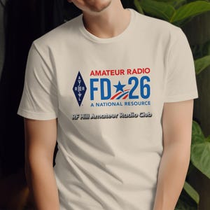 Può includere: Una t-shirt a maniche corte color crema con una grafica che riporta il testo AMATEUR RADIO FD 26 A NATIONAL RESOURCE, un logo ARRL e la scritta RF Hill Amateur Radio Club in nero sotto il design principale di colore blu e rosso.