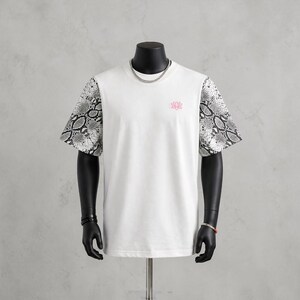 Könnte beinhalten: Weißes T-Shirt mit kurzen Ärmeln mit Schlangenmuster und einem kleinen rosa Lotusblüten-Emblem. Das Shirt wird auf einer Schaufensterpuppe mit einer silbernen Halskette und Armbändern präsentiert.