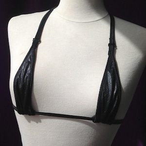 Könnte beinhalten: Ein schwarzes Bikini-Oberteil mit einem dezenten Prägemuster auf dem Stoff. Das Kleidungsstück besteht aus zwei dreieckigen Körbchen, dünnen verstellbaren Trägern mit Kunststoffschiebern und einem schmalen horizontalen Band.