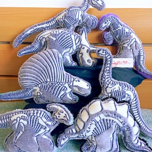 Op de afbeelding: Een verzameling van zeven pluche dinosaurusknuffels gemaakt van grijze, pluizige stof met witte, gezeefdrukte skeletontwerpen. Elk zacht speelgoed heeft een eigen prehistorische vorm, waaronder langnek- en stekelsoorten, met een wit label Lil-Sime.