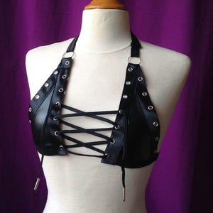 Puede incluir: Un top negro estilo halter con cordones negros cruzados en la parte delantera. La prenda está confeccionada en un material liso y semibrillante, decorada con ojales metálicos de color plateado a lo largo de los tirantes y paneles centrales.