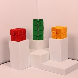 May include: Three colorful fidget cubes in red, green, and yellow are displayed on white pedestals. The red and yellow cubes are in a cube shape, while the green one is elongated. The background is a light pink.