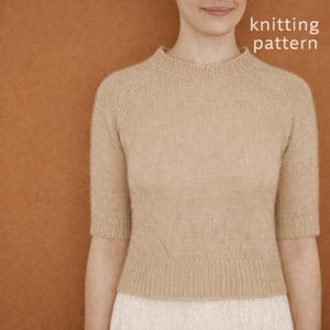 May include: A beige knitted top with short sleeves and a high neckline. The top is made of a soft, textured yarn and has a ribbed hem and cuffs. The words "knitting pattern" are in the upper right corner.