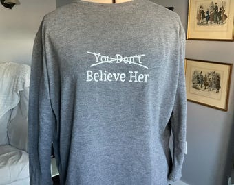 Könnte beinhalten: Ein graues, langärmeliges Shirt auf einer Schneiderpuppe. Das Shirt ist mit dem weißen Schriftzug "You Don't Believe Her" bedruckt, wobei die Worte "You Don't" durch eine horizontale Linie durchgestrichen sind. Der Code "A-072" ist rosa.
