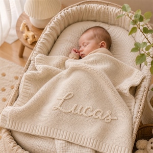 Op de afbeelding: Een slapende baby ligt in een geweven wieg, bedekt met een cr&egrave;mekleurige gebreide deken. De deken heeft de naam "Lucas" in een scriptlettertype. De wieg is gevoerd met een zachte, neutrale stof.