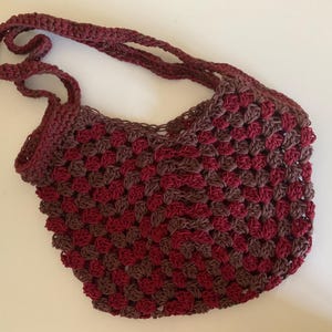 May include: A handmade crochet bag in a granny square pattern. The bag features a combination of burgundy and brown yarn, with a matching burgundy strap. The bag is designed for carrying items and has a shoulder strap.