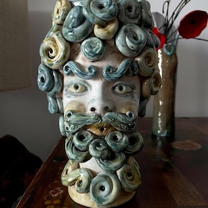 Puede incluir: Un jarrón de cerámica con forma de rostro barbudo con pelo rizado y bigote. La cara es blanca con ojos amarillos y detalles azules en el pelo y la barba. Un jarrón con flores rojas está en el fondo.