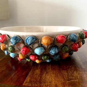 Puede incluir: Cuenco de cerámica decorativo con interior crema y exterior texturizado y colorido. El borde está adornado con un patrón de formas redondeadas multicolores en rojo, azul, naranja y verde. Ideal para decoración.