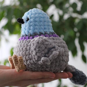 以下が含まれることがあります： 青、グレー、紫の色合いの手作りのぬいぐるみハト。鳥は黒いクチバシと小さなベージュの足を持っています。柔らかく、質感のある素材が鳥に抱きしめたくなるような外観を与えています。ユニークな手作りのアイテムです。