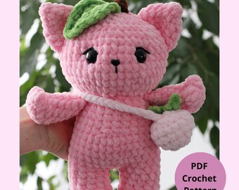May include: A handmade pink crochet cat with a green leaf and brown stem on its head, and a white strap holding a pink fruit. The cat has black eyes and a small black nose. The text "Peachy the cat" is at the top.