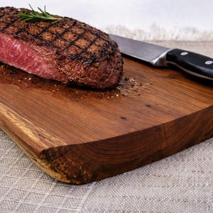 Könnte beinhalten: Ein gegrilltes Steak mit einem Kreuzmuster, das auf einem Holzschneidebrett liegt. Ein Rosmarinzweig liegt auf dem Steak. Ein Messer mit schwarzem Griff liegt neben dem Steak.