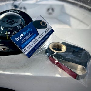 May include: A black and silver Ritchie compass with a clear dome sits on a white surface. A blue and white DockReady card and a black and red tool are also visible. The card reads "2-IN-1 BILGE PLUG HOLDER & WRENCH TOOL".