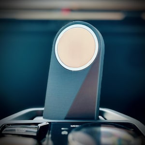 May include: A dark gray rectangular device with a beige circular element at the top. The device has a geometric design with a brown triangle on the side. The device is set in a car's console.