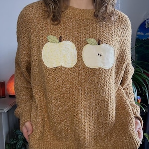 Könnte beinhalten: Ein hellbrauner Strickpullover mit zwei aufgenähten Apfelmotiven. Die Äpfel sind cremefarben mit grünen Blättern und braunen Stielen. Der Pullover hat einen Rundhalsausschnitt und lange Ärmel. Das Strickmuster ist über dem gesamten Kleidungsstück sichtbar.