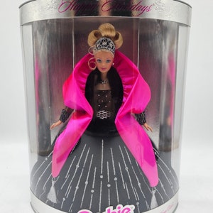 May include: A Barbie doll wearing a black gown with silver accents and a pink fur-trimmed coat. The doll is in a clear plastic box with a silver background and the words "Happy Holidays" printed on the top. The box also has the Barbie logo and the words "Precious Gift" printed on the bottom.