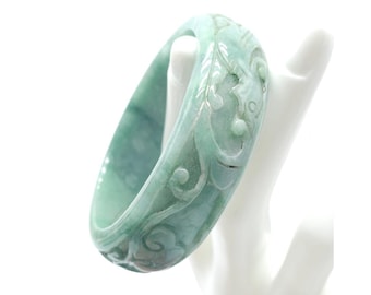 May include: A light green jade bangle bracelet with intricate carved floral and scroll designs. The jade bracelet is displayed on a white hand-shaped stand, showcasing its smooth, polished surface and detailed carvings.