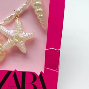 K&ouml;nnte beinhalten: Nahaufnahme einer Halskette mit Muschel- und Seestern-Anh&auml;ngern. Die Anh&auml;nger sind irisierend wei&szlig; und h&auml;ngen an einer wei&szlig;en Perlenkette. Die Halskette wird auf einem rosa Hintergrund pr&auml;sentiert, mit einer leuchtend rosa Schachtel.