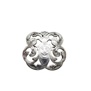 Peut inclure: Une broche argentée avec un design complexe mettant en scène un visage stylisé au centre.