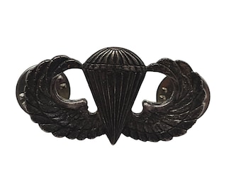 May include: A silver-toned military paratrooper badge with wings and a parachute. The badge has a pin back.