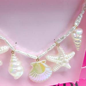 K&ouml;nnte beinhalten: Halskette mit Charms aus irisierenden wei&szlig;en Muscheln und Seesternen. Die Halskette hat eine Kette aus wei&szlig;en Perlen mit goldfarbenen Akzenten. Die Charms umfassen eine Muschel, eine Jakobsmuschel und einen Seestern. Die Halskette wird auf rosa Hintergrund pr&auml;sentiert.