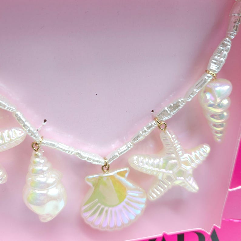 K&ouml;nnte beinhalten: Halskette mit Charms aus irisierenden wei&szlig;en Muscheln und Seesternen. Die Halskette hat eine Kette aus wei&szlig;en Perlen mit goldfarbenen Akzenten. Die Charms umfassen eine Muschel, eine Jakobsmuschel und einen Seestern. Die Halskette wird auf rosa Hintergrund pr&auml;sentiert.