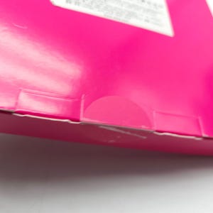 K&ouml;nnte beinhalten: Nahaufnahme einer leuchtend rosa Pappschachtel mit einem teilweise sichtbaren wei&szlig;en Etikett. Die Schachtel hat eine gefaltete Kante und einen halbkreisf&ouml;rmigen Ausschnitt. Die Oberfl&auml;che ist gl&auml;nzend und reflektiert Licht.